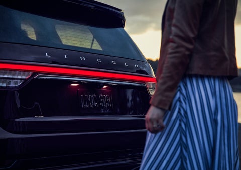 A person is shown near the rear of a 2024 Lincoln Aviator® SUV as the Lincoln Embrace illuminates the rear lights | Beck Lincoln in Palatka FL