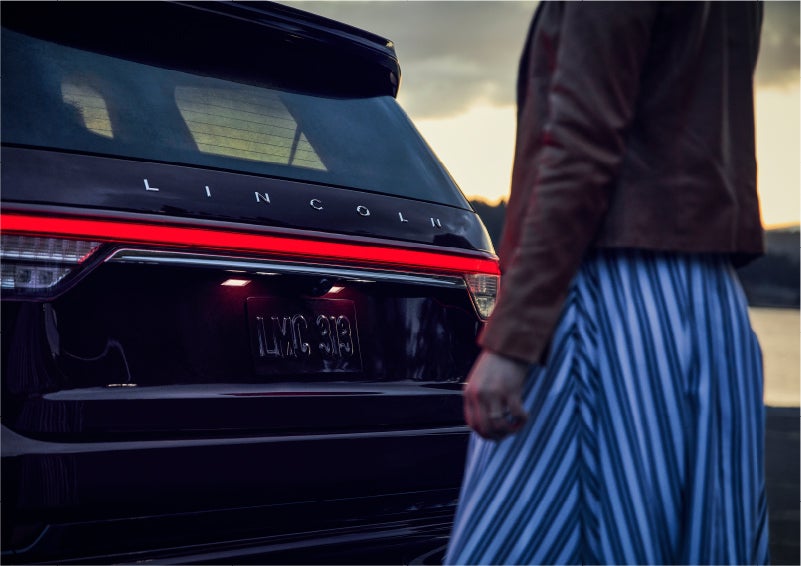 A person is shown near the rear of a 2023 Lincoln Aviator® SUV as the Lincoln Embrace illuminates the rear lights | Beck Lincoln in Palatka FL